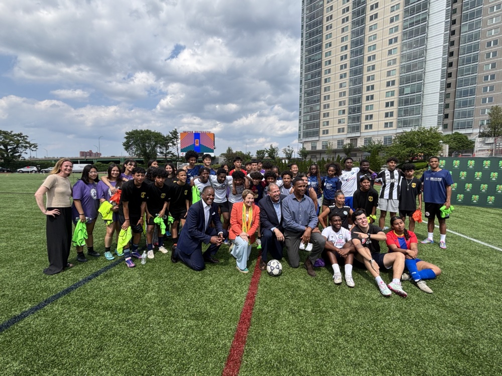 A large group of high school-aged youth, coaches, and partners gather on a soccer field in front of a tall apartment building and a digital scoreboard during the announcement of the Boston Host Committee and U.S. Soccer Foundation partnership. Everyone is smiling and posing together, some holding neon green shirts, with a soccer ball placed at the center of the group. The event takes place on a sunny day with a partly cloudy sky.